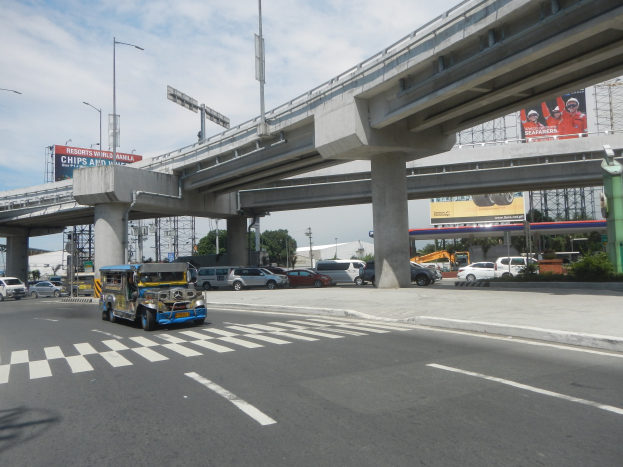 Eine verkehrsreiche Hauptstraße mit Fahrzeugen, einer Zebrastreifen, einer Überführung mit Pfeilern, Laternen, Schildern mit Text und Bildern, Bäumen, Gebäuden und einem bewölkten Himmel im Hintergrund.
