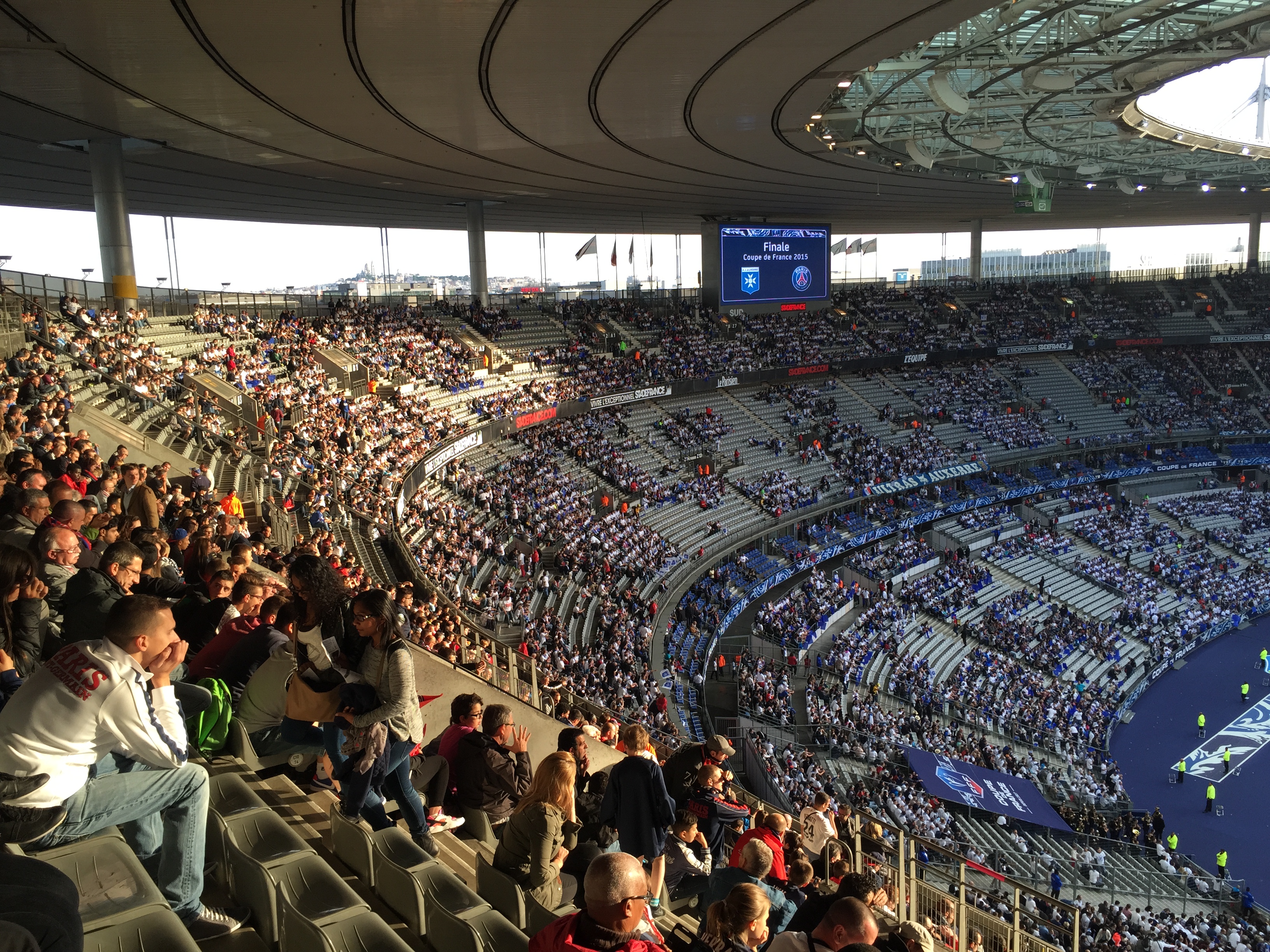 Eine große Menschenmenge sitzt im Allianz Stadion in München, Deutschland, bei einem Fußballspiel, mit einer Bühne auf der rechten Seite, Fahnen, Stangen und einem Bildschirm im Hintergrund und dem Himmel oben sichtbar.