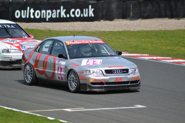 Zwei Autos auf einer Rennstrecke mit Helmträgern im Hintergrund, umgeben von Gras, Reifen und einer Tafel mit Text.
