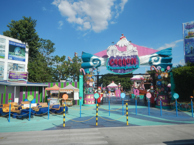 Karussell am Parkeingang mit Straße, Absperrung, Buden, Fahnen, Bäumen und bewölktem Himmel im Hintergrund.