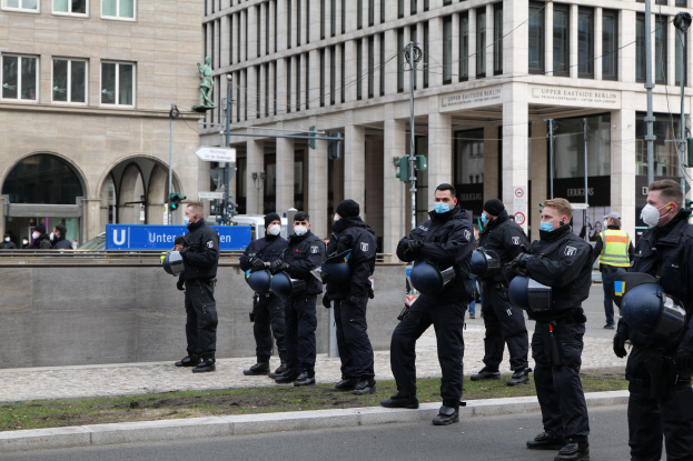 Eine Gruppe von Polizisten in schwarzen Uniformen und Masken steht vor einem Gebäude mit Glasfenstern und Säulen in Berlin, Deutschland, einige halten Helme, mit Laternen, Verkehrsampeln, Texttafeln, einer Statue, anderen Menschen und einem grasbewachsenen Boden im Hintergrund.