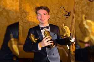 Ein Mann im Smoking lächelt und hält eine Oscar-Statue in der Hand, mit einer Wand im Hintergrund und einem Wasserzeichen auf dem Bild.
