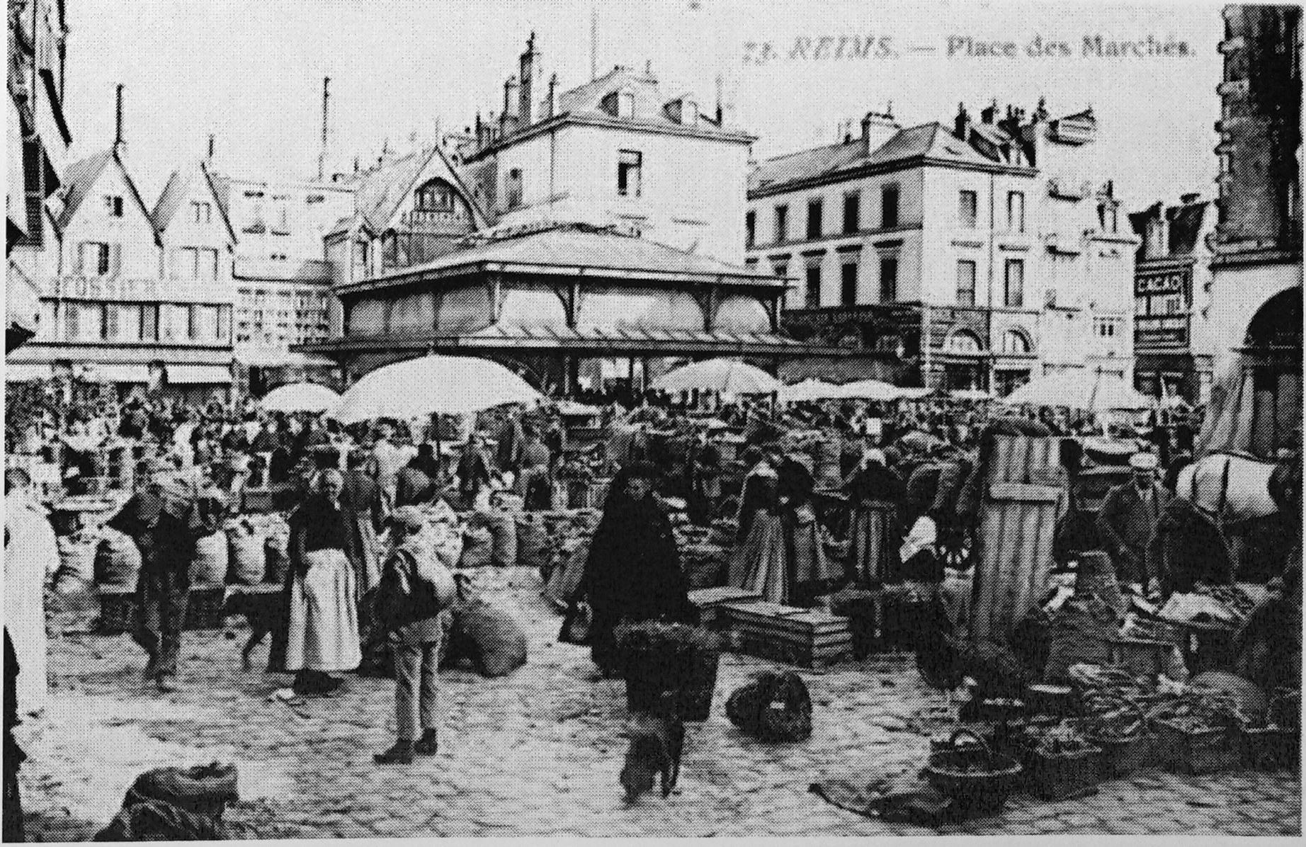 Schwarzes und weißes Foto eines belebten Stadtmarkts mit Menschen, Schirmen, verstreuten Körben und Gebäuden im Hintergrund.