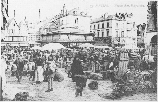 Schwarzes und weißes Foto eines belebten Stadtmarkts mit Menschen, Schirmen, verstreuten Körben und Gebäuden im Hintergrund.