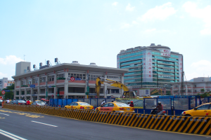 Eine belebte Stadtstraße mit zahlreichen Fahrzeugen, Fußgängern, hohen Gebäuden, einem Kran, Verkehrsampeln, Schildern, Bäumen und einem bewölkten Himmel.