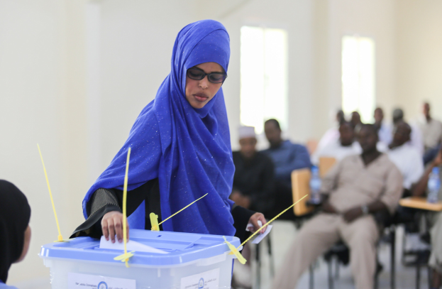 Eine Frau mit blauem Kopftuch gibt ihre Stimme in einer Wahllokal ab, während andere in der Nähe sitzen.