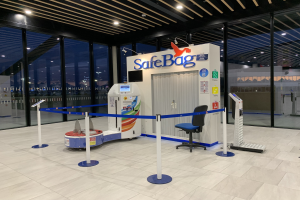 Sicherheitskontrolle am Flughafen mit Stuhl, Absperrpollern, Maschinen, Bildschirm, bedruckter Tafel, Glaswänden, Deckenleuchten und sichtbarem Boden.