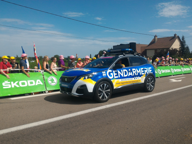 Polizeiauto fährt an einer Menge vorbei, die Fahnen schwenkt und ein Banner auf dem Fahrzeug mit den Worten "Gendarmerie de la Tour de France" am unteren Rand.