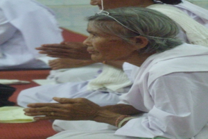 Eine ältere Frau liegt im Krankenhausbett und hat ihre Hände gefaltet, sie sieht besorgt aus mit geschlossenen Augen und Kopf auf den Händen, umgeben von ein paar Menschen im Hintergrund.