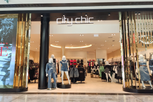 Ein stylischer Modeboutique in einem Einkaufszentrum mit Schaufensterpuppen in der neuesten Mode, Kleider an Bügeln, einer Säule, einem Schild, Deckenleuchten und einem Fliesenboden.