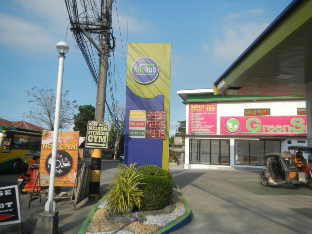 Tankstelle mit einem "Green's"-Schild, Fahrzeuge auf der Straße, umliegende Pflanzen, Schilder, Pfosten, Lampen, Drähte, Gebäude, Bäume und Wolken am Himmel.