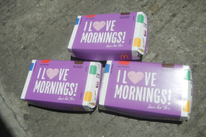 Drei buntfarbene McDonald's I Love Mornings-Schachteln mit dem McDonald's-Logo und weißer Schrift auf einem hellblauen Himmel mit weißen Wolken.