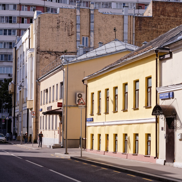 Eine Stadtstraße mit einem gelben Gebäude, fahrenden Fahrzeugen, Fußgängern auf dem Gehweg, Straßenschildern, hohen Gebäuden, Bäumen und einem klaren blauen Himmel.