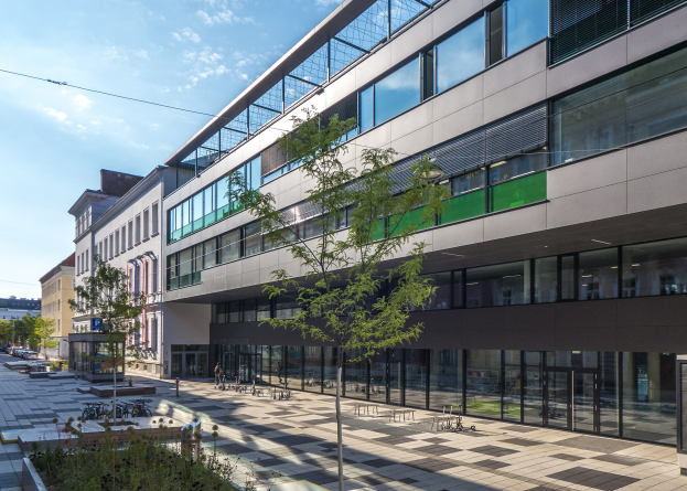 Großes modernes Gebäude mit vielen Fenstern, umgeben von Bäumen und Fahrzeugen, mit Menschen in der Nähe und bewölktem Himmel im Hintergrund; als das neue Hauptquartier des Deutschen Instituts für Technologie in Berlin bezeichnet.