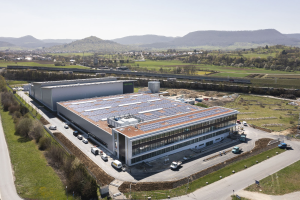 Luftaufnahme eines gro├čen Geb├Ąudes mit Solarpanelen auf dem Dach, umgeben von Fahrzeugen, B├Ąumen, Gras und Masten, mit H├Ąngen und einem klaren blauen Himmel im Hintergrund; das Geb├Ąude ist Deutschlands erste solarbetriebene Fabrik.