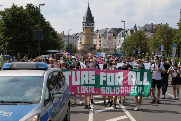 Eine große Gruppe von Menschen marschiert mit einem Banner, auf dem "Lokale Solidarität" steht, eine Straße entlang, während ein Polizeiwagen daneben parkt, Bäume und Gebäude im Hintergrund unter einem bewölkten Himmel.