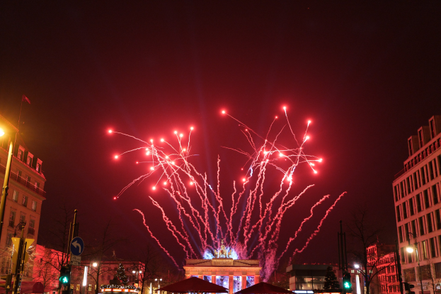 Eine belebte Stadtstraße bei Nacht während des Silvesterfeuerwerks in Berlin mit Gebäuden, Bäumen, Laternenmasten, Ampeln, Schildern, Zelten, Menschen und einem prächtigen Feuerwerk am Himmel.