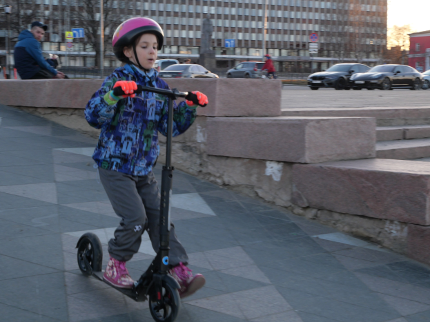 Ein junger Junge in Helm und Handschuhen fährt ein Scooter eine Treppe hinunter auf einem Gehweg mit Fahrzeugen, Menschen, Bäumen, Pfählen, Brettern, Gebäuden und einem klaren blauen Himmel im Hintergrund.
