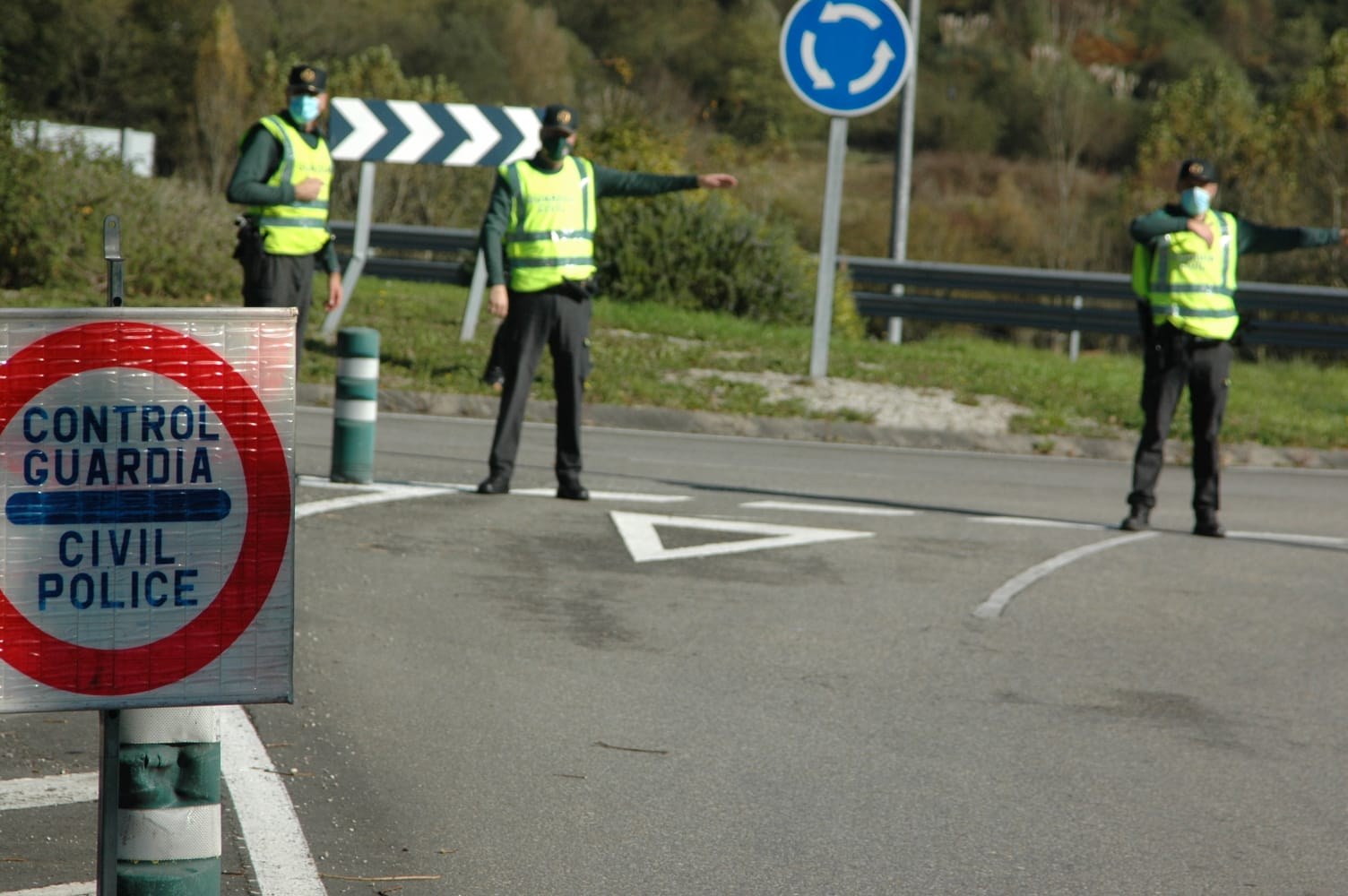 Drei Polizeibeamte in grünen Jacken und schwarzen Hosen stehen an einer Straßenrand bei Bäumen, einem Geländer, einem Verkehrsschild und einem Schuh.
