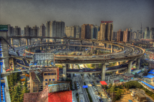Eine Stadtansicht mit einer zentralen Ringstraße mit Fahrzeugen, einer unteren Straße mit Autos und Fahrrädern, zahlreichen Hochhäusern im Hintergrund, einem Himmel darüber, Bäumen auf der linken unteren Seite und kleinen Häusern, die über die Stadt verteilt sind.