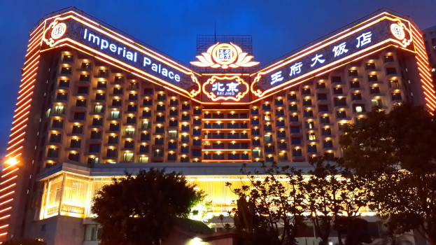 Das Kaiserpalast-Hotel in Peking, China, ein großes mehrstöckiges Gebäude mit zahlreichen Fenstern und beleuchteter Beschriftung, vor einem klaren blauen Himmel mit Bäumen im Vordergrund.