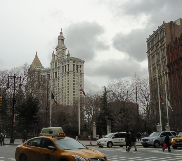 Gelbes Taxi fährt auf einer von Hochhäusern gesäumten Stadtstraße, Fußgänger auf dem Gehweg, Verkehrskegel, Laternenmasten, Fahnenmasten, Bäume und eine bewölkte Kulisse im Hintergrund.
