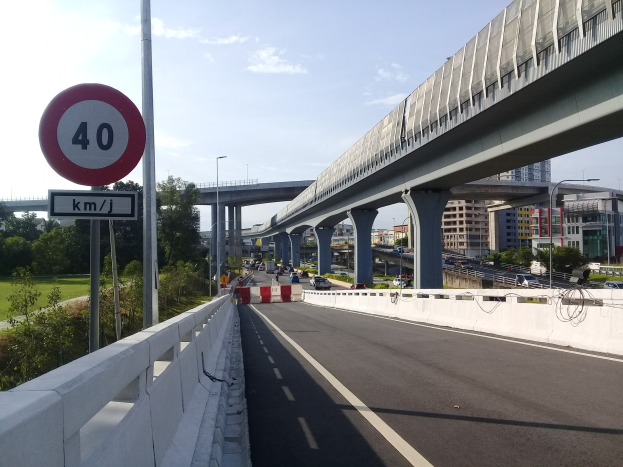 Highway with speed limit sign, vehicles, bridge pillars, light poles, trees, buildings, and cloudy sky in the background.