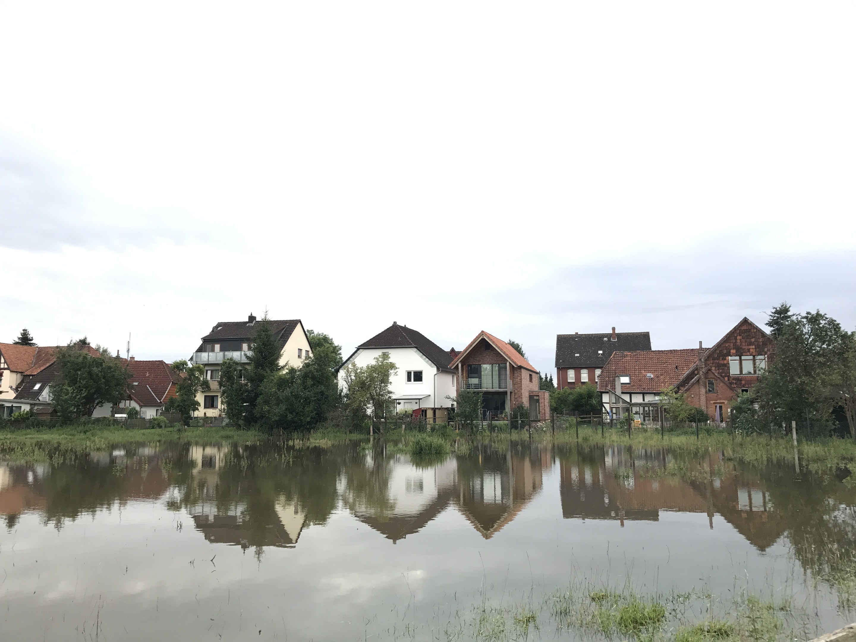 Überflutete Straße mit teilweise unter Wasser stehenden Häusern, umgeben von Gras, Pflanzen, Bäumen und Strommasten, unter einem sichtbaren Himmel.