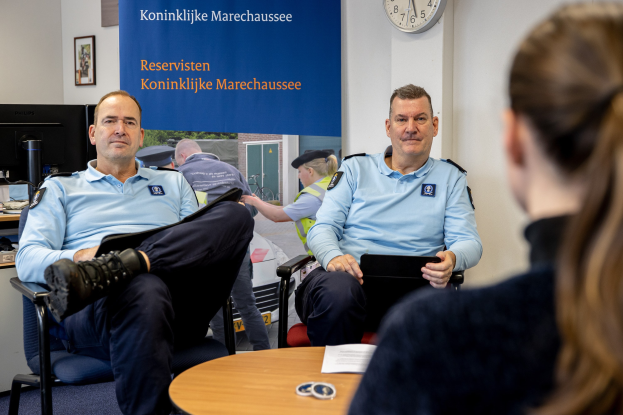 Zwei Polizeibeamte sitzen an einem Tisch mit Papieren und Gegenständen, sprechen mit einer Frau, mit einer Fahne, Uhr und Foto Rahmen im Hintergrund und einem weiteren Tisch mit Monitor und Tastatur links.