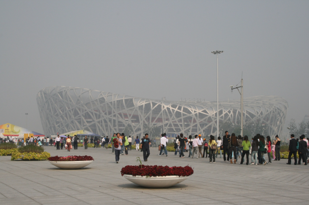 Olympiastadion in Peking mit Menschen, die herumlaufen, Blumenpflanzen im Vordergrund, Laternenmasten, Bäume und einen klaren blauen Himmel im Hintergrund.