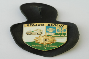Ein schildförmiges Polizeibadge mit "Polizei Berlin" in schwarzer fettgedruckter Schrift auf einem blauen Hintergrund mit einem weißen Stern und Rand, das auf einer Oberfläche platziert ist.