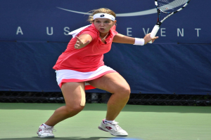 Eine Frau in einem pinken Shirt und weißen Schuhen, die ein Headband trägt, spielt Badminton auf einem Court und hält einen Schläger, wobei ein Banner im Hintergrund sichtbar ist.