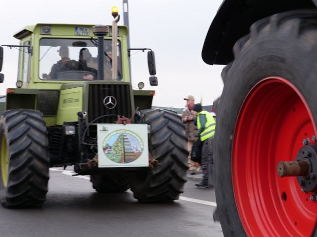 Ein grüner Traktor mit einem roten Rad fährt eine Straße entlang, umgeben von Menschen in Jacken und Mützen, mit Bäumen und einem klaren blauen Himmel im Hintergrund.
