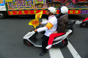 Zwei Personen mit Helmen fahren auf einem Motorroller, eine hält eine gelbe Flagge, im Hintergrund ist ein Fahrzeug zu sehen.