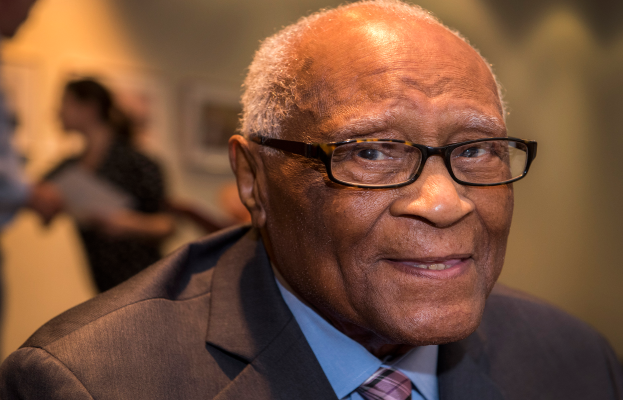 Bill Cosby, ein älterer Mann in Anzug, Krawatte und Brille, lächelt in die Kamera mit einem unscharfen Hintergrund.