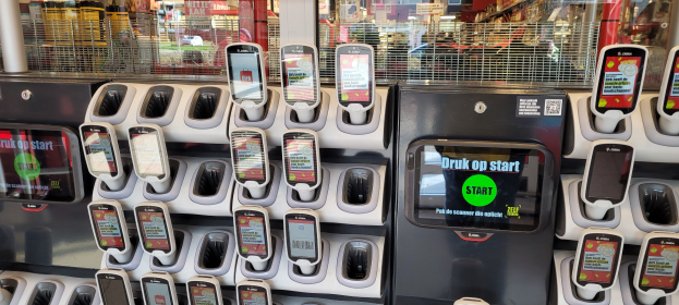 Ein Verkaufsautomat mit mehreren Handys darauf, vor einem Hintergrund voller Menschen, Geschäfte, Schilder und Lichter.