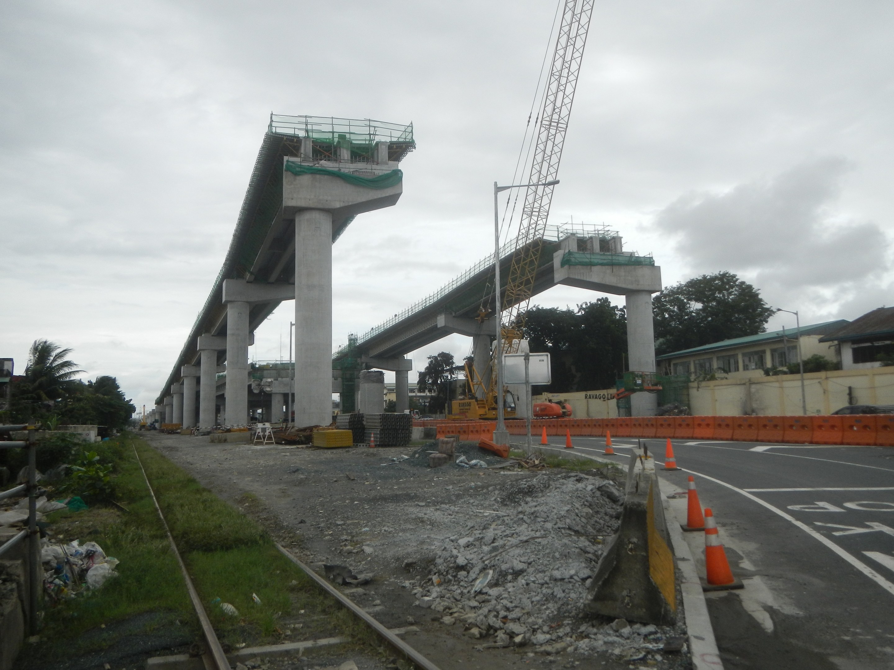 Baustelle mit Brücke im Hintergrund, Straße mit Absperrkegeln auf der rechten Seite, Bahnschiene auf der linken Seite, verstreute Steine und Gras, Bäume und Gebäude auf beiden Seiten und ein bewölkter Himmel.