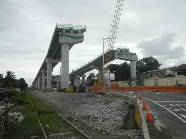 Baustelle mit Brücke im Hintergrund, Straße mit Absperrkegeln auf der rechten Seite, Bahnschiene auf der linken Seite, verstreute Steine und Gras, Bäume und Gebäude auf beiden Seiten und ein bewölkter Himmel.