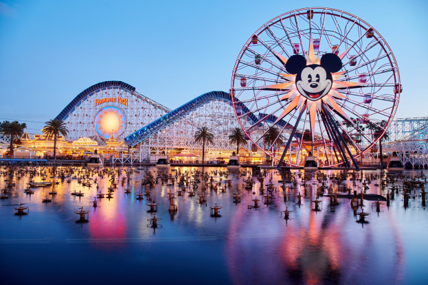 Leuchtendes Disneyland California Adventure-Szenario bei Sonnenuntergang mit einem riesigen Mickey-Maus-Rad im Vordergrund, einer Achterbahn im Hintergrund und Booten auf dem Wasser, mit einem bunten Himmel und silhouettenartigen Bäumen.