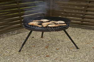 Grill mit Fleisch darauf auf einem Kiesgelände, umgeben von einem Holzzaun und Bäumen, mit Rauch, der aus dem Grill aufsteigt.