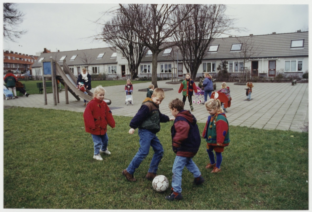 Kinder spielen Fußball auf Rasen in einem Park mit Bäumen, Gebäuden, Bänken und einer Rutsche im Hintergrund unter einem sichtbaren Himmel.