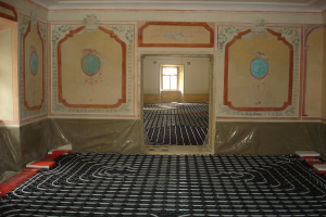 Ein Raum mit einer Fußbodenheizung in der Mitte, umgeben von Wänden und einem Fenster im Hintergrund, mit verschiedenen Gegenständen auf dem Boden, die mit dem Heizsystem in Verbindung stehen.