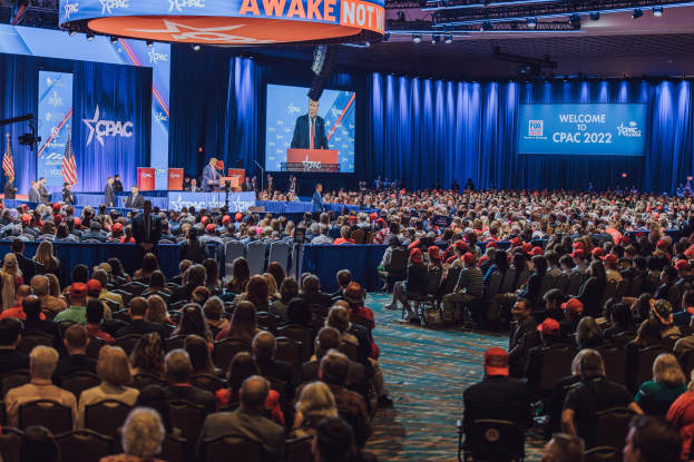 Eine große Menschenmenge sitzt auf Stühlen, die einer Bühne zugewandt sind, auf der ein paar Menschen auf einem Podium stehen, mit anderen, die links davon stehen, blauen Vorhängen und einem Bildschirm im Hintergrund und einer orangenen Tafel oben, auf der "CPAC 2020" und "25. Oktober" steht.