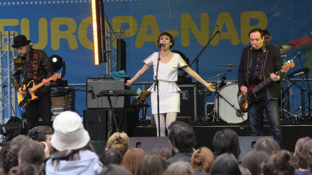 Vier Musiker treten auf der Bühne auf: Eine Frau in einem weißen Kleid singt in der Mitte, während zwei Männer links und rechts Gitarren spielen, mit einer sichtbaren Menge unten und Lautsprechern im Hintergrund an der Wand montiert.