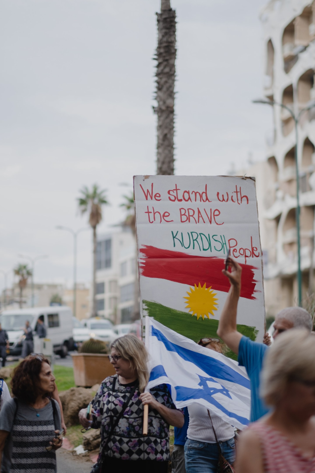Eine Gruppe von Menschen geht eine Straße entlang und hält ein Schild hoch, auf dem steht "Wir stehen zu den mutigen kurdischen Menschen", mit einem auf der Seite geparkten Van, Straßenlaternen, Bäumen, Gebäuden und dem Himmel im Hintergrund.