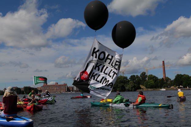 Gruppe von Menschen in Kajaks auf dem Wasser mit einem "Kohle Kill Klima"-Schild, Paddel in der Hand, vor dem Hintergrund von Bäumen, Gebäuden, Kränen und blauem Himmel.