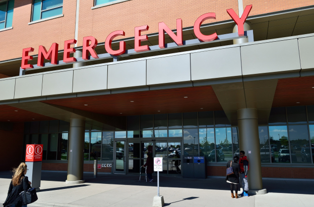 Gruppe von Menschen, die auf ein Notaufnahmelokal mit Glaswänden, Säulen, einer Namensschild, Hinweistafeln und einem Abfalleimer zusteuern.