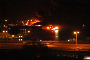 Großbrand in einer Stadt bei Nacht mit Straßenlaternen, einem Baum im Vordergrund, einem dunklen Himmel und naher Wasserfläche.