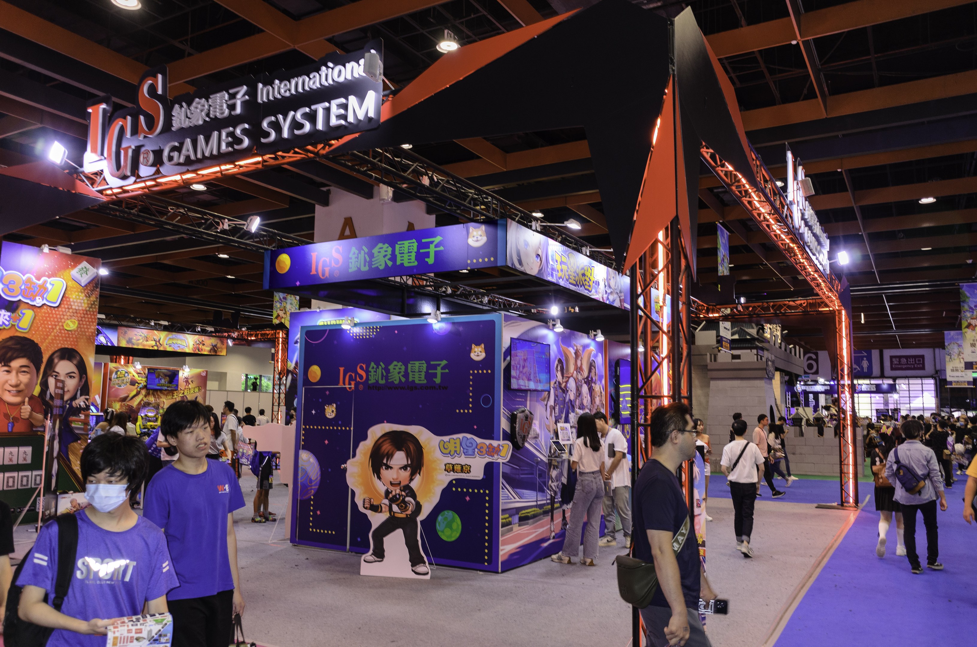 Eine Gruppe von Teilnehmern durchquert einen Veranstaltungsraum auf einer Spielemesse in China, einige tragen Taschen und halten Gegenstände, mit Displaytafeln, Deckenbeleuchtung und Teppichboden.