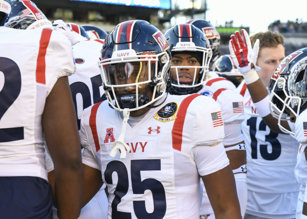 Gruppe von Männern in Football-Uniformen auf einem Feld mit Helmen und Handschuhen, mit einer unscharfen Menge, einem Scoreboard mit der Aufschrift "Auburn vs Virginia Tech - 11/28/15 College Football Pick, Odds, and Prediction" und Himmel im Hintergrund.